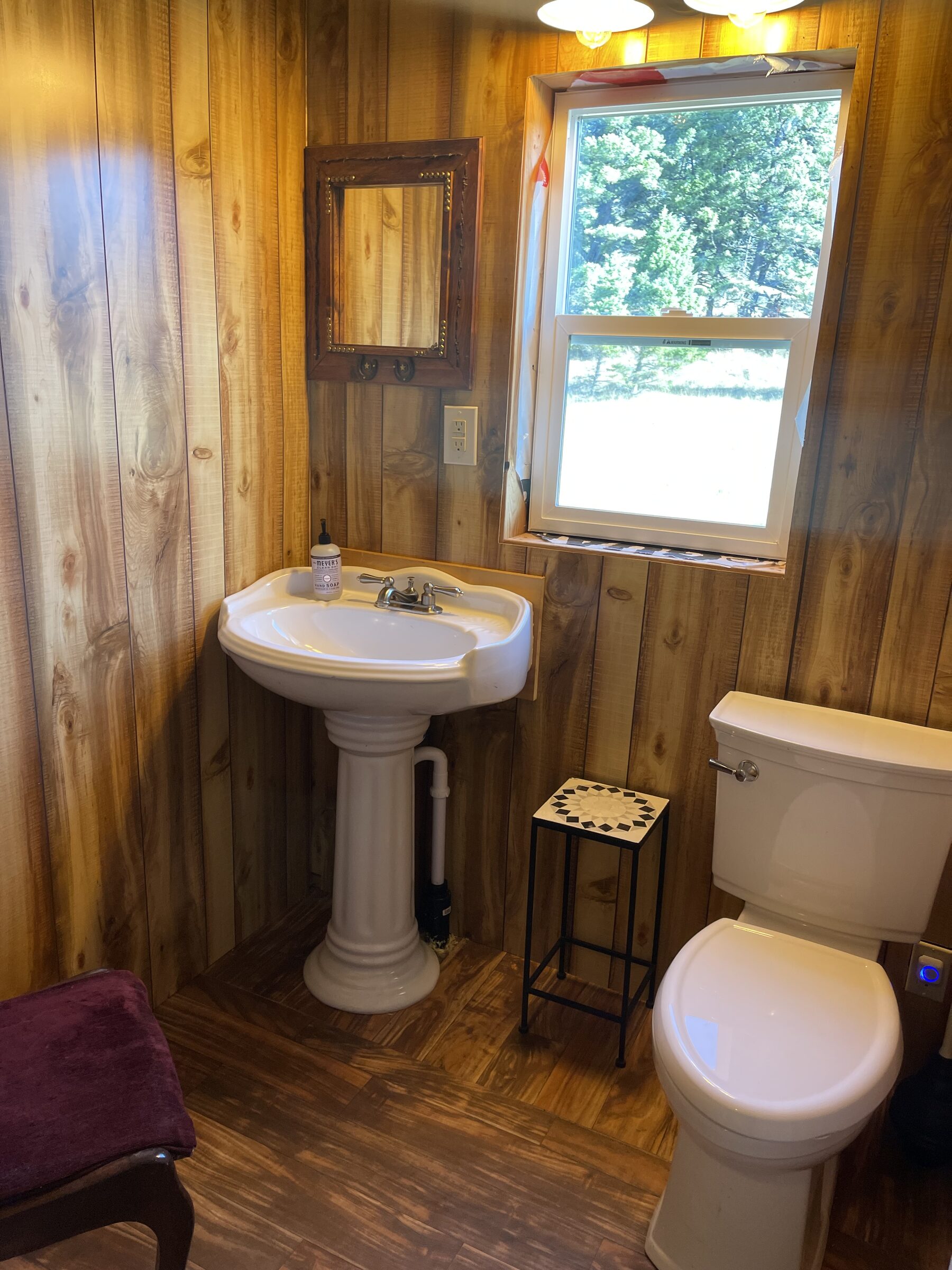 The pedestal-sink bathroom in the Whiskey Ridge Boarding House at Sweetwater Ranch — a white pedestal sink, a framed mirror, and a window to the pines