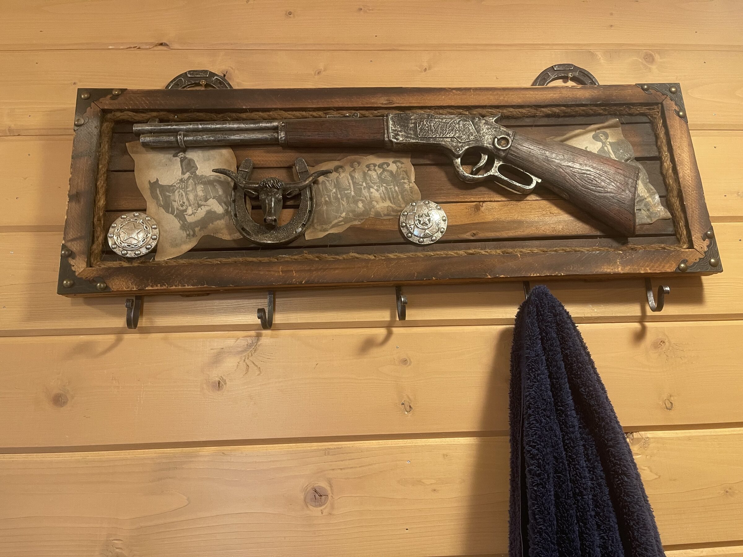 A Western shadowbox on the knotty-pine wall of the Main Cabin at Sweetwater Ranch — a lever-action, a longhorn, conchos, and old cowboy photographs, hung above a row of towel hooks