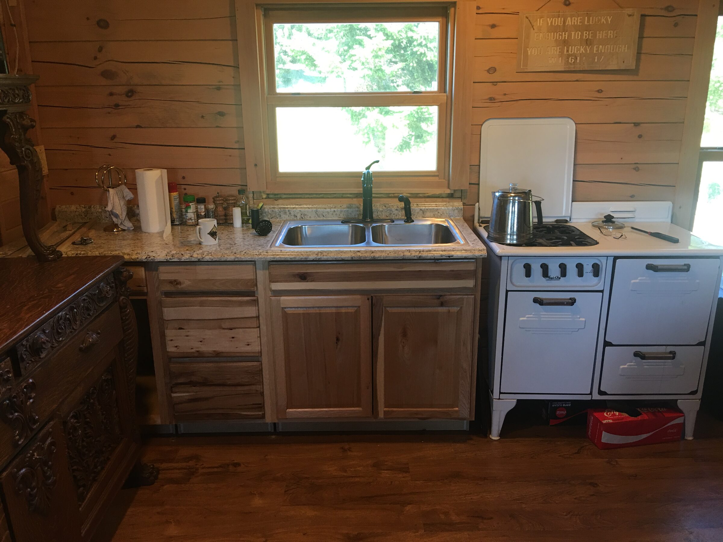 The cabin kitchen at Sweetwater Ranch — counters and a double sink beneath the window, a vintage enamel range, and a hand-carved antique sideboard against knotty-pine walls