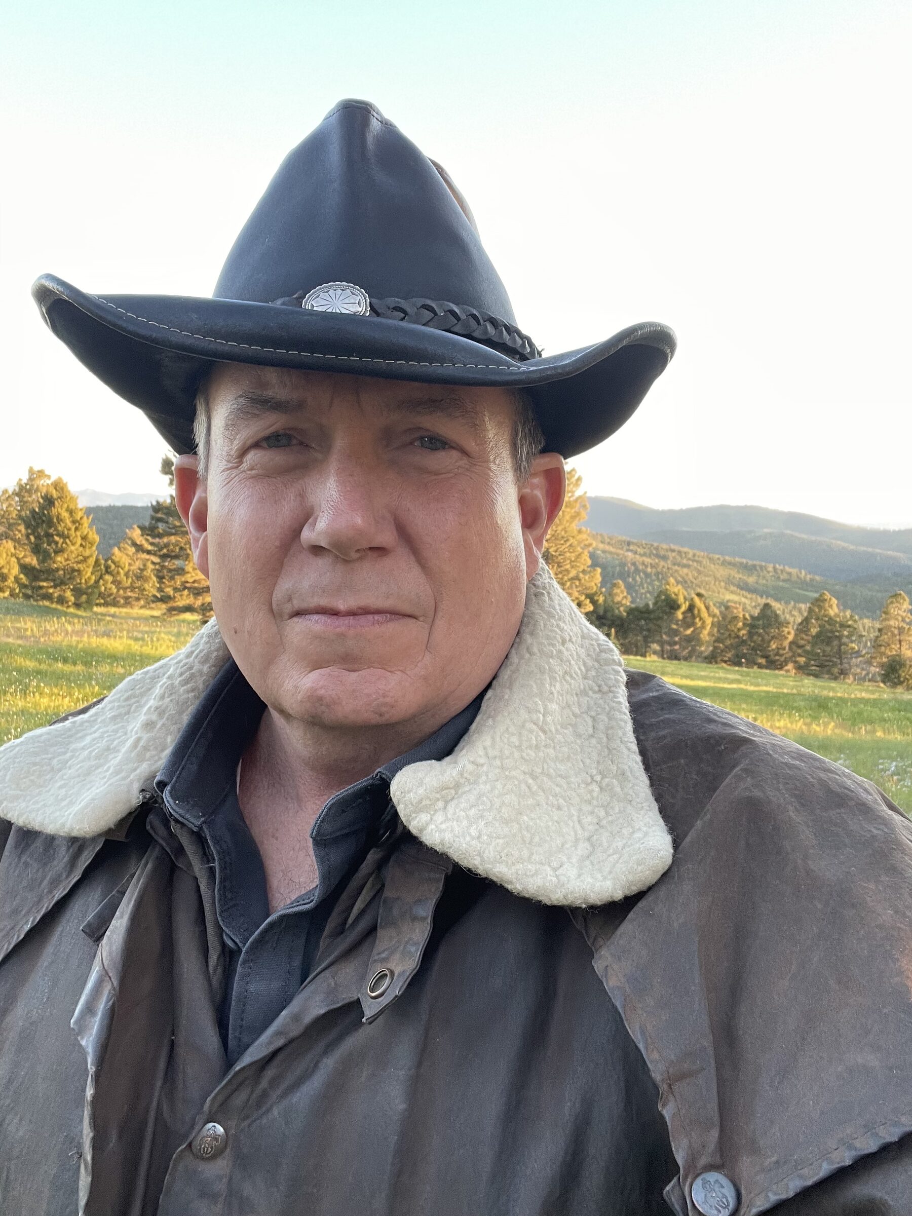 Terry L. Fossum in a leather outback hat and oilskin duster at golden hour, the Montana range glowing behind him at Sweetwater Ranch