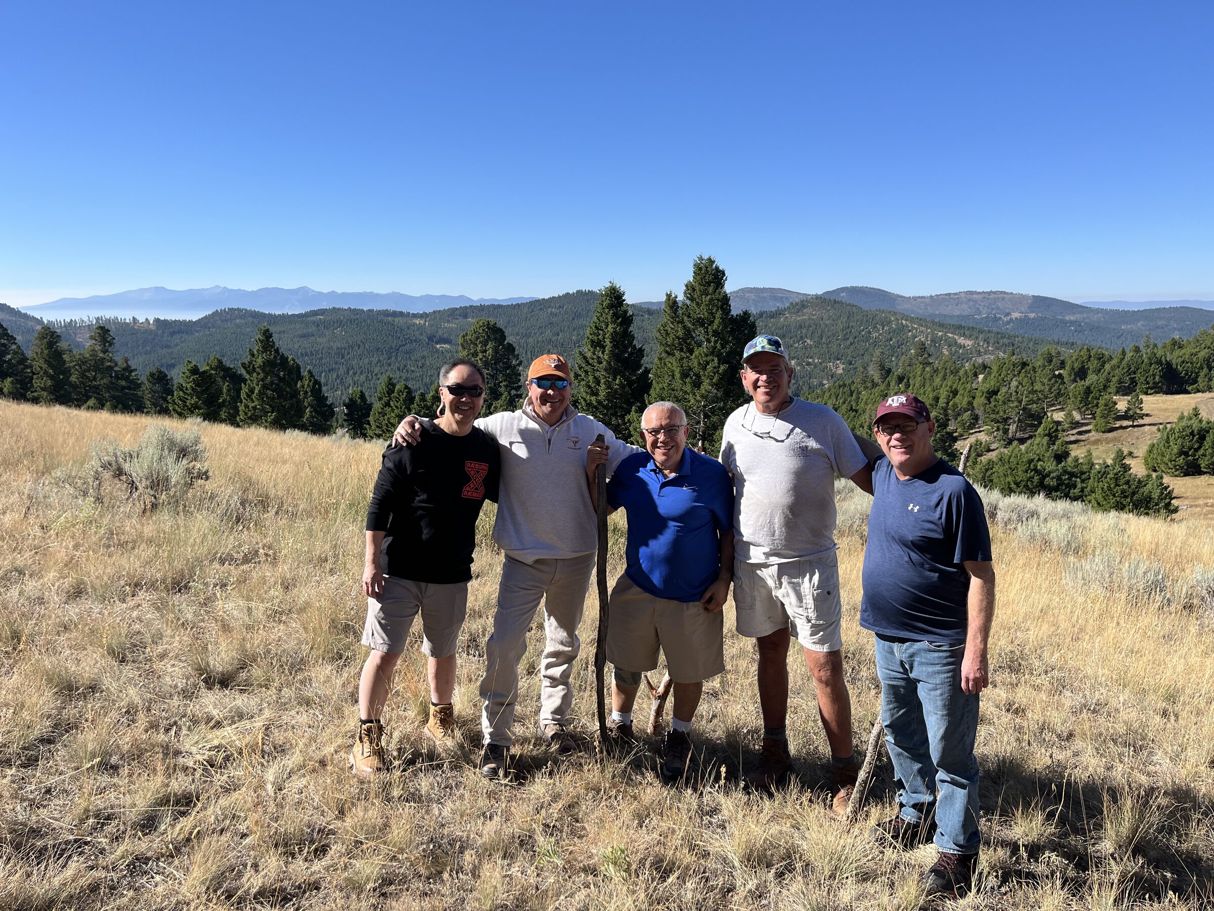 Five friends arm in arm on the golden meadow at Sweetwater Ranch, layered Montana mountain ranges behind them