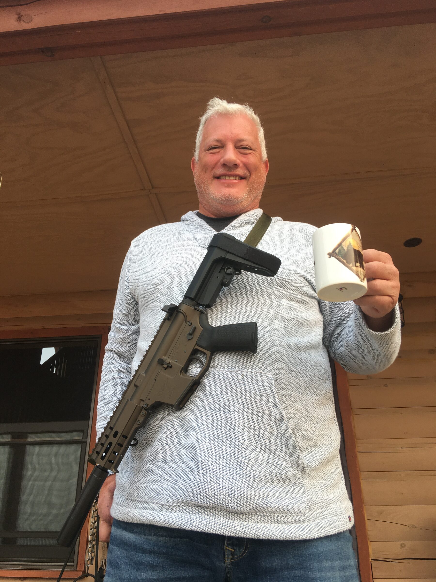 A guest on the cabin porch at Sweetwater Ranch, a coffee mug in one hand and a rifle slung over his shoulder, grinning