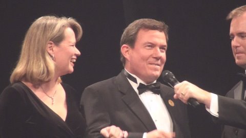 Terry L. Fossum being interviewed on camera at a gala, a microphone held to him, his wife Michelle at his side