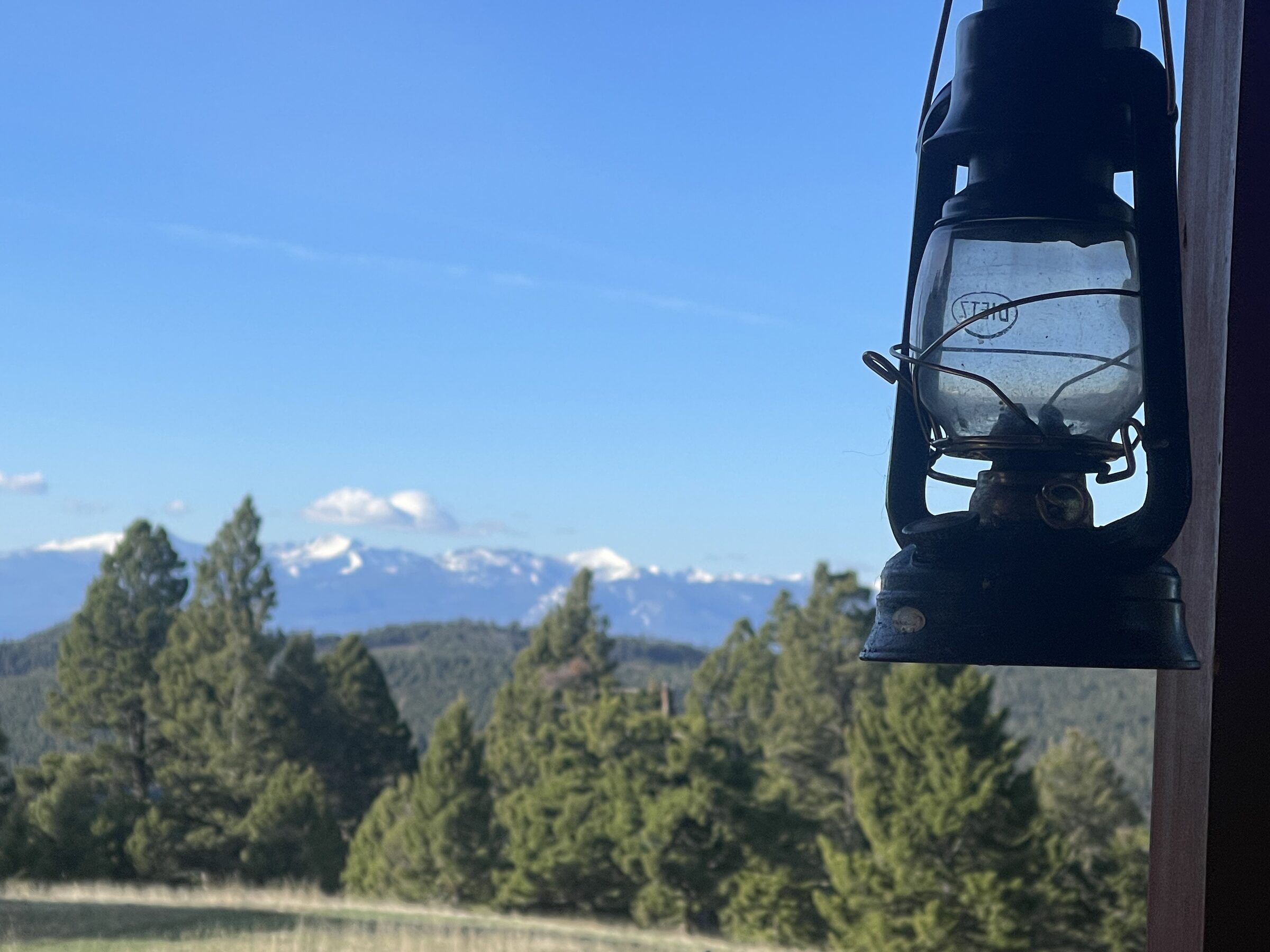 An antique kerosene lantern hanging on the cabin porch at Sweetwater Ranch, the snow-capped range beyond the pines