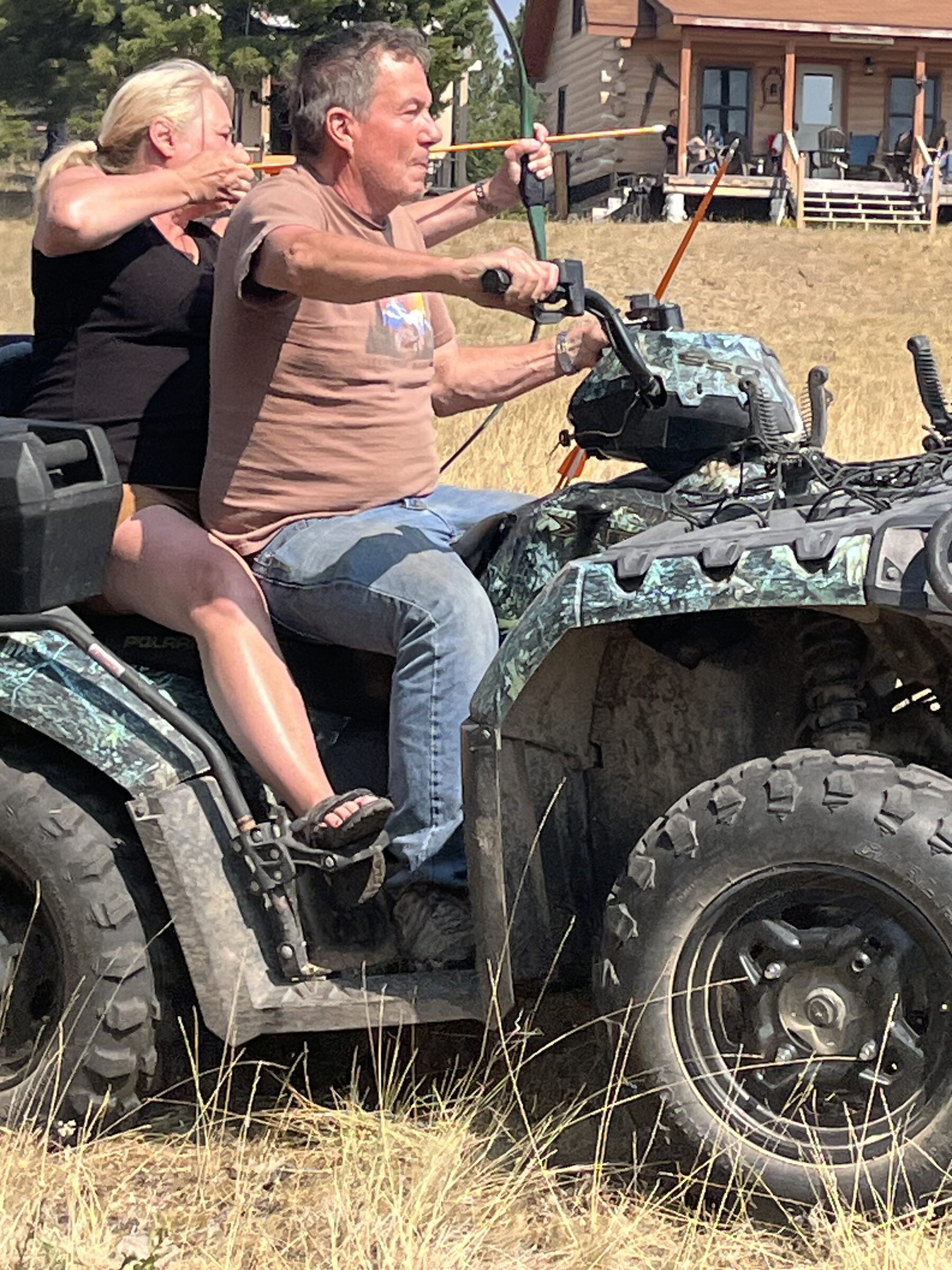 A couple on a quad at Sweetwater Ranch — one driving while the other draws a bow for target practice, the cabin behind them