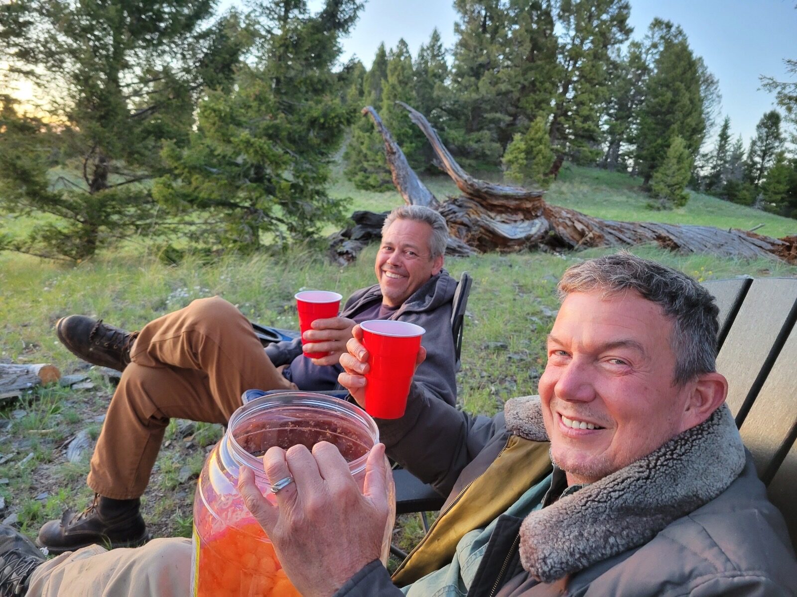 Two friends in camp chairs at golden hour on the mountain at Sweetwater Ranch, red cups raised, a jar of sangria between them