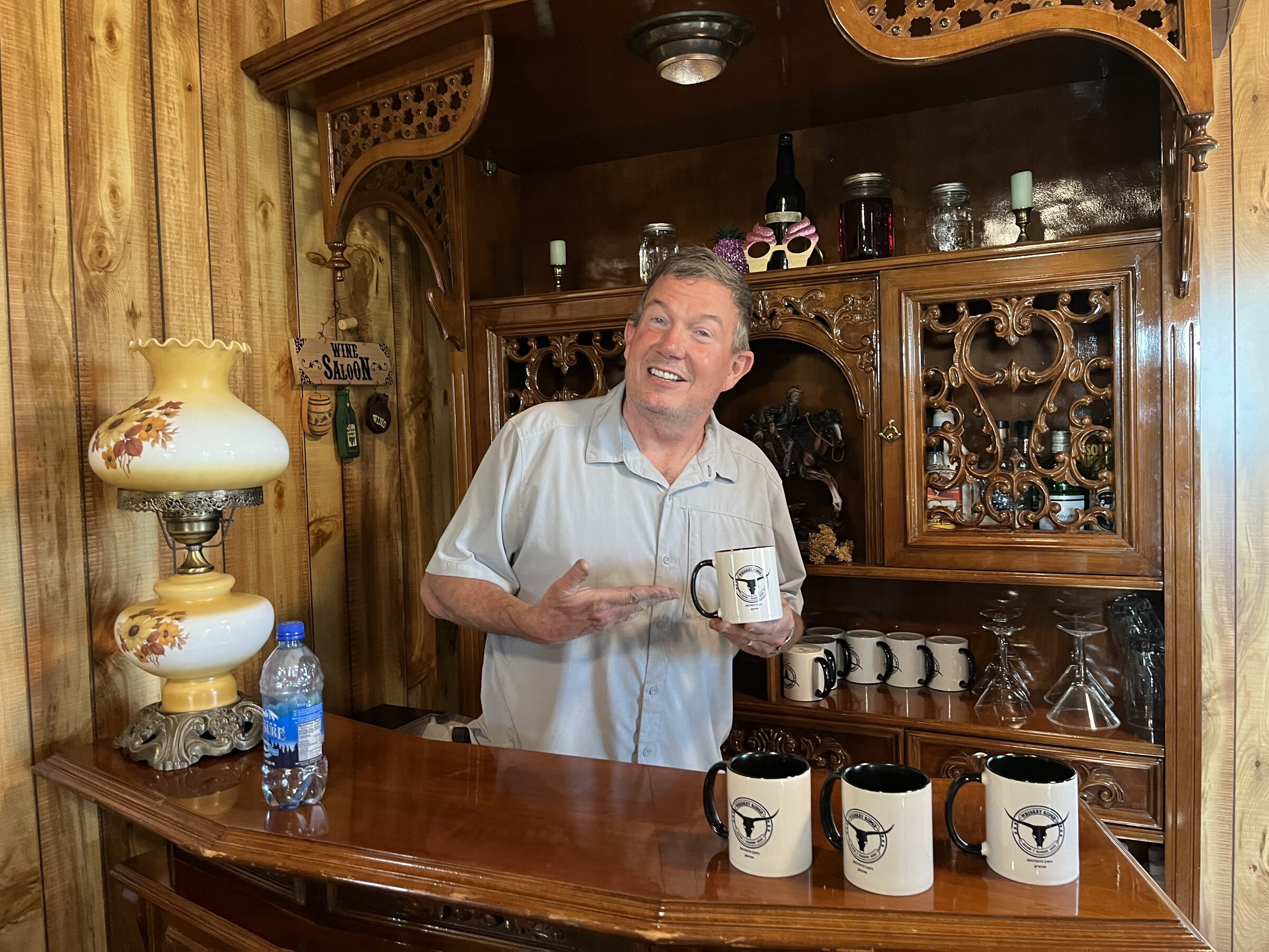 The owner standing behind the carved bar at the Whiskey Ridge Saloon at Sweetwater Ranch, with the Whiskey Ridge mugs and a Wine Saloon sign
