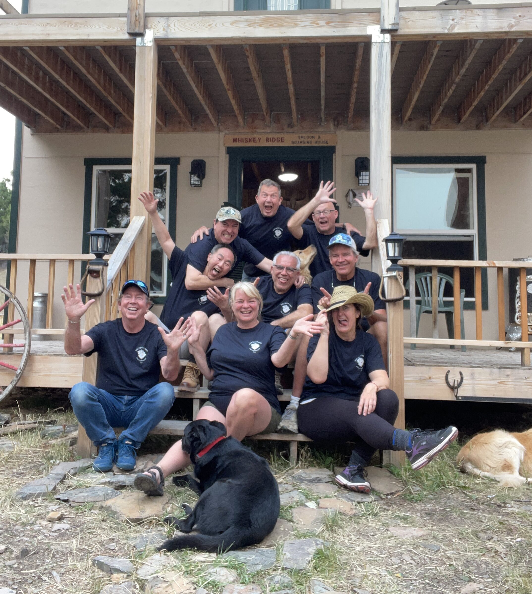 A crowd of friends in matching shirts laughing on the steps of the Whiskey Ridge Saloon & Boarding House at Sweetwater Ranch, two dogs at their feet