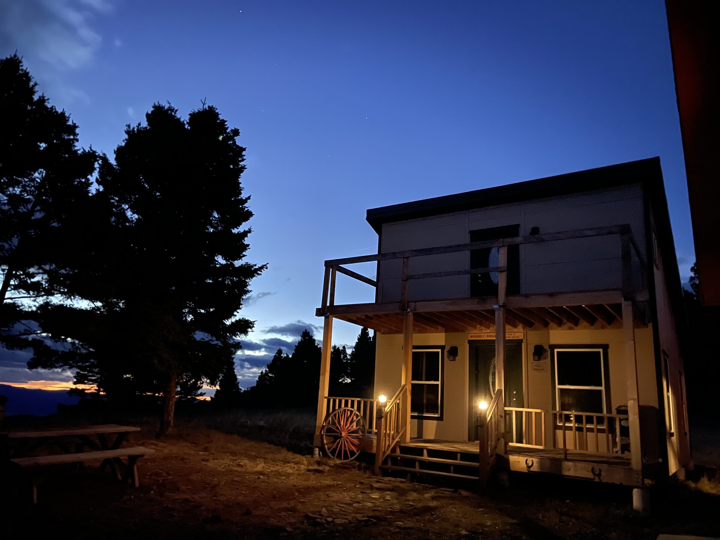The Whiskey Ridge Saloon & Boarding House at Sweetwater Ranch lit up at dusk — a porch lamp glowing, a wagon wheel on the rail, the last of the sunset behind the pines